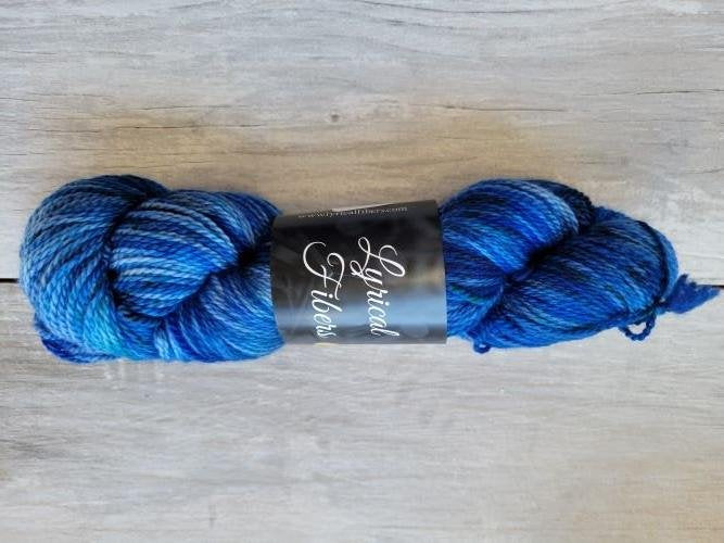 A skein of blue variegated yarn labeled "Lyrical Fibers" is placed on a light wooden surface. The yarn shows a mix of dark and light blue shades with some purple tones.
