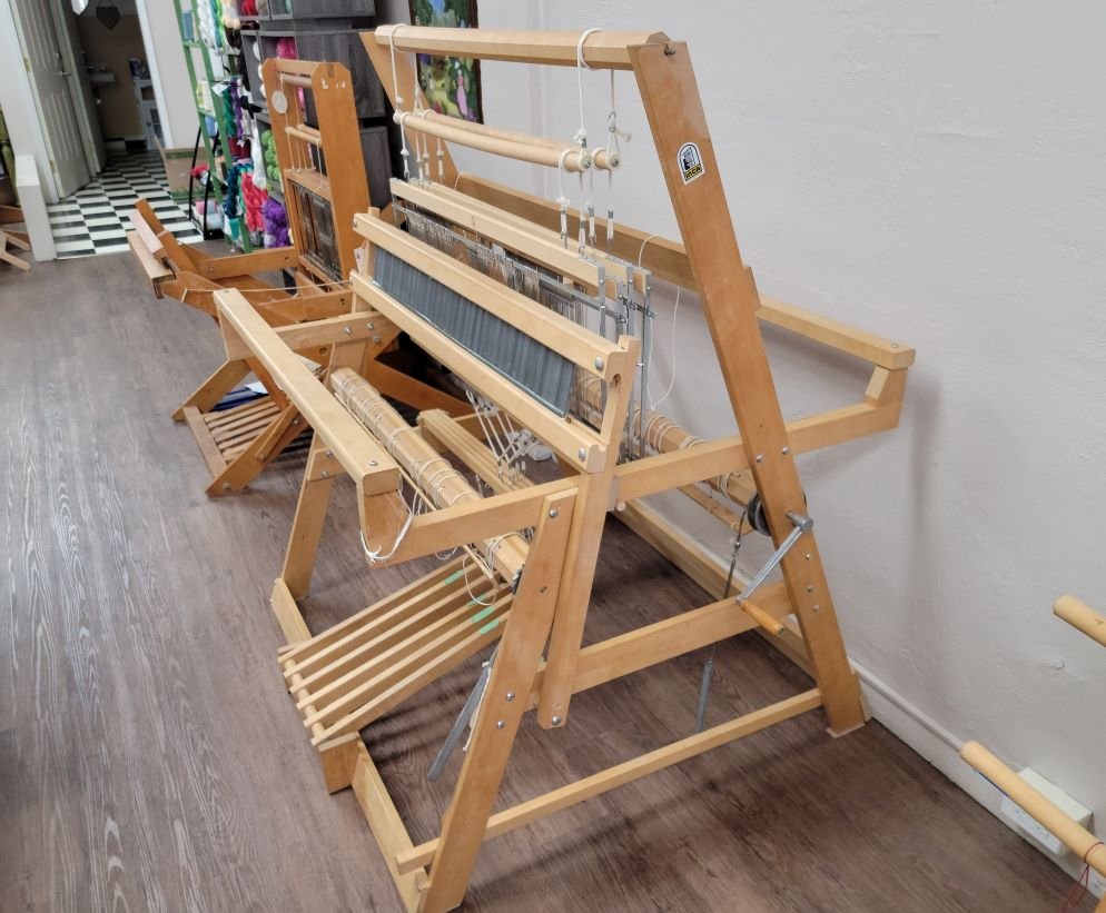A wooden weaving loom is positioned in a room with a hard floor. The loom appears to be well-constructed with several shafts and treadles. In the background, another similar loom can be seen, along with a checkered floor pattern.