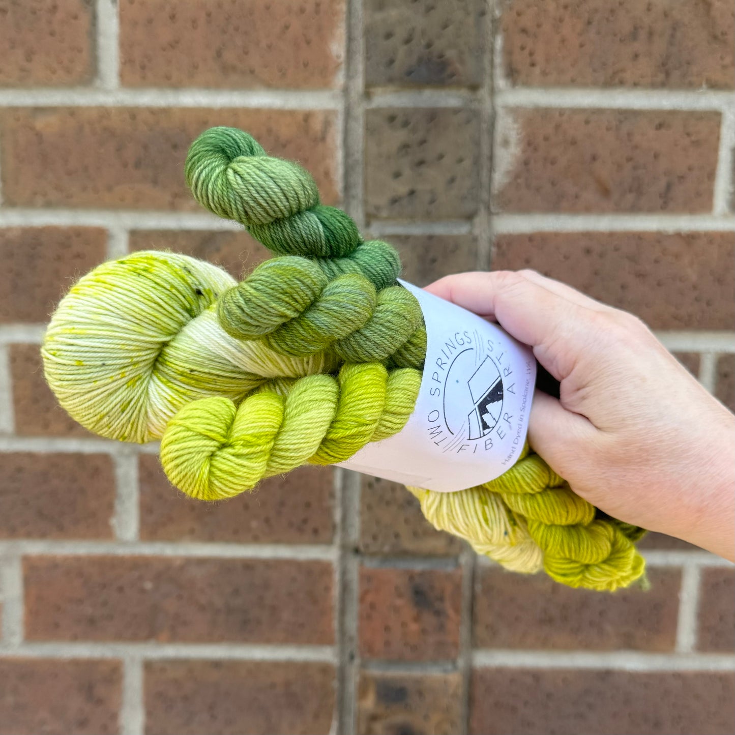 A basket filled with various colorful skeins of yarn, including shades of red, purple, green, blue, and multicolored. Each skein has a label with brand information. The basket is held outside against a brick wall background, partially in sunlight.