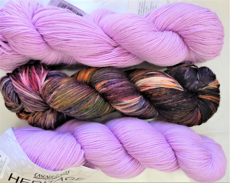 Three skeins of yarn are displayed. The top and bottom skeins are solid light purple, while the middle skein is a mix of dark brown, light brown, purple, pink, and green colors. All skeins are from Cascade Yarns' Heritage series.