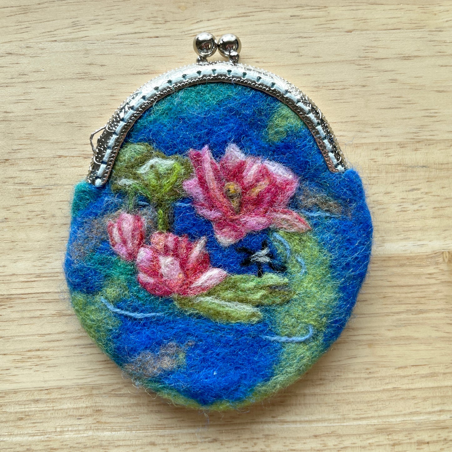 Round, wet felted coin purse with floral design of lotus in a pond on a wooden surface