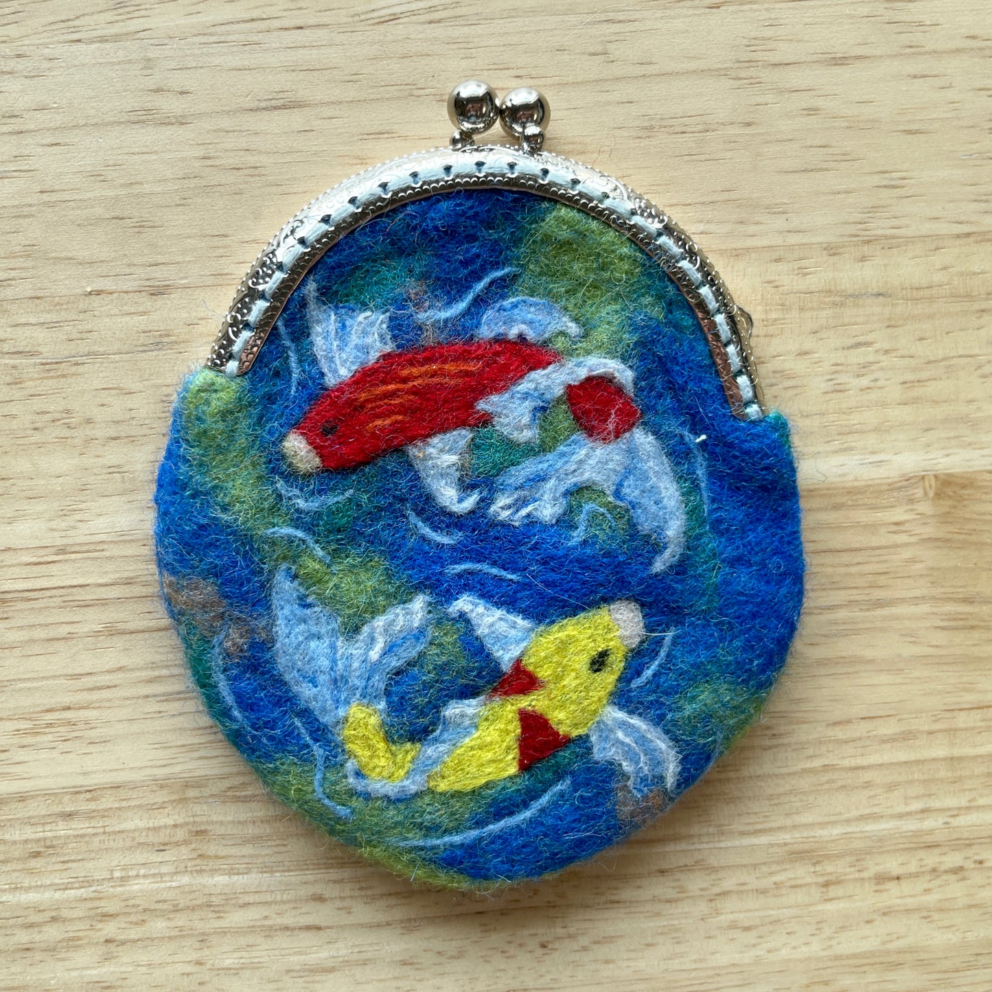Round, wet felted coin purse with embroidered design of fish on a wooden surface