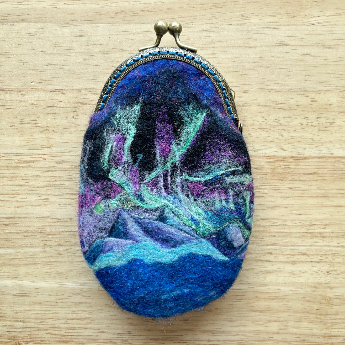 Oval-shaped, wet felted coin purse with a colorful design of the aurora borealis on a wooden surface