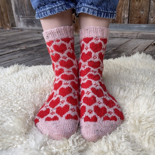 Two feet wearing knitted socks with a red heart pattern. The socks have a pink background, and the toes are pointed down. The person is standing on a white fluffy surface, and the background includes wooden flooring and a wooden door.