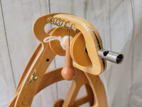 A close-up image of a wooden spinning wheel. The image focuses on the orifice and surrounding components, including hooks and a distaff, highlighting the detailed construction and craftsmanship of the spinning wheel. A loop of thread hangs from the orifice.