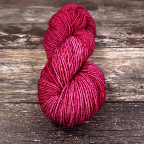 A skein of red yarn is coiled in a loose spiral and placed on a wooden surface. The yarn has various shades of red, ranging from deep crimson to lighter reds, giving it a vibrant appearance. The wooden surface is rustic with visible grain and textures.