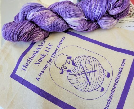 A skein of purple and white variegated yarn lies on a beige tote bag. The bag reads "The Hook & Needle Nook, LLC A Haven for Fiber Artists" and features a cartoon sheep wrapped in yarn holding knitting needles.okset.