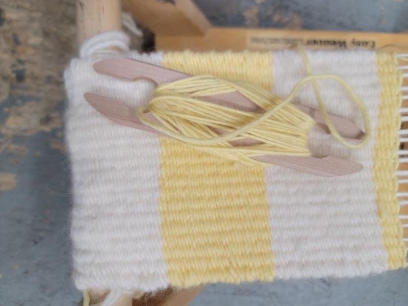 A close-up image of a partially woven textile on a small loom. The textile features alternating white and yellow horizontal stripes. Two wooden shuttles with yellow yarn are resting on the loom.