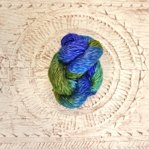 A skein of blue and green yarn is placed on a textured white surface with an intricate circular pattern. The yarn is tightly twisted, showcasing a blend of vivid blue and green hues.