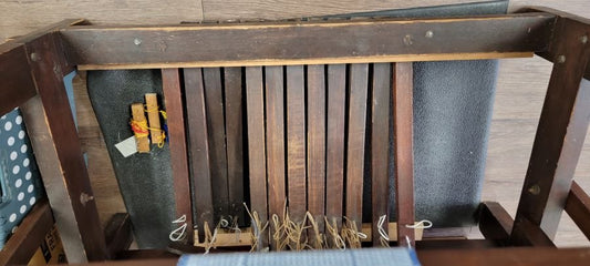 A view of the underside of a wooden chair showing support beams, springs, and a piece of fabric. There are a few small items, including a bundle of yellowish strings and a clear plastic bag with multicolored items inside, resting on the slats.