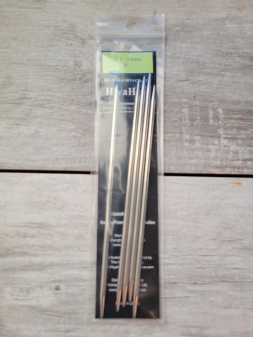 A plastic package containing five double-pointed knitting needles with pointed tips is placed on a wooden surface. A green and dark label inside the package reads "HiyaHiya Stainless Steel..." indicating the brand and material of the knitting needles.