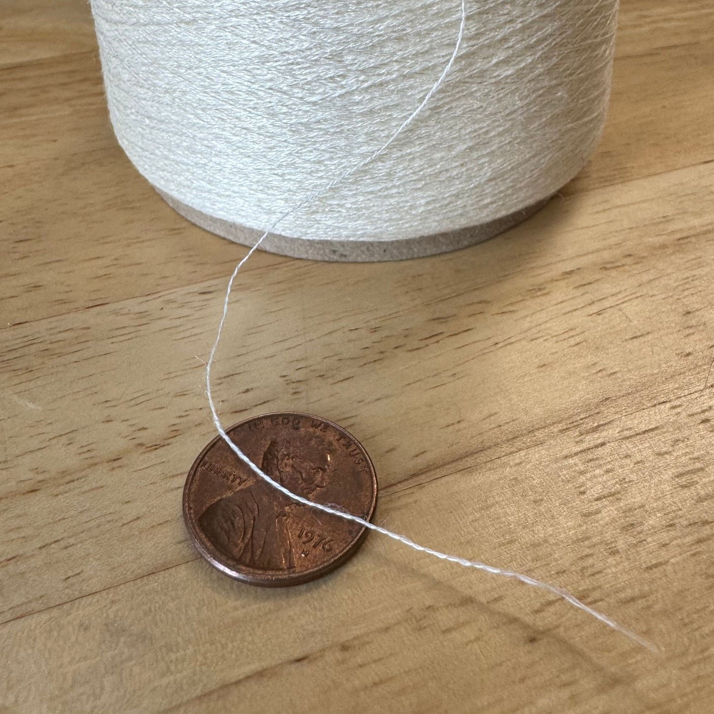 A close-up image shows a brown U.S. penny next to a beige spool of thread. The penny is dated 1976 and there is a strand of white thread on the wooden surface near it. The spool and the penny provide a sense of scale for the thickness of the thread.