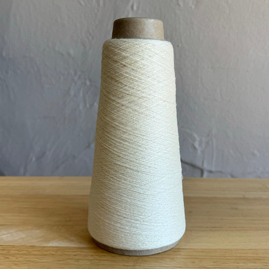 A close-up image shows a brown U.S. penny next to a beige spool of thread. The penny is dated 1976 and there is a strand of white thread on the wooden surface near it. The spool and the penny provide a sense of scale for the thickness of the thread.