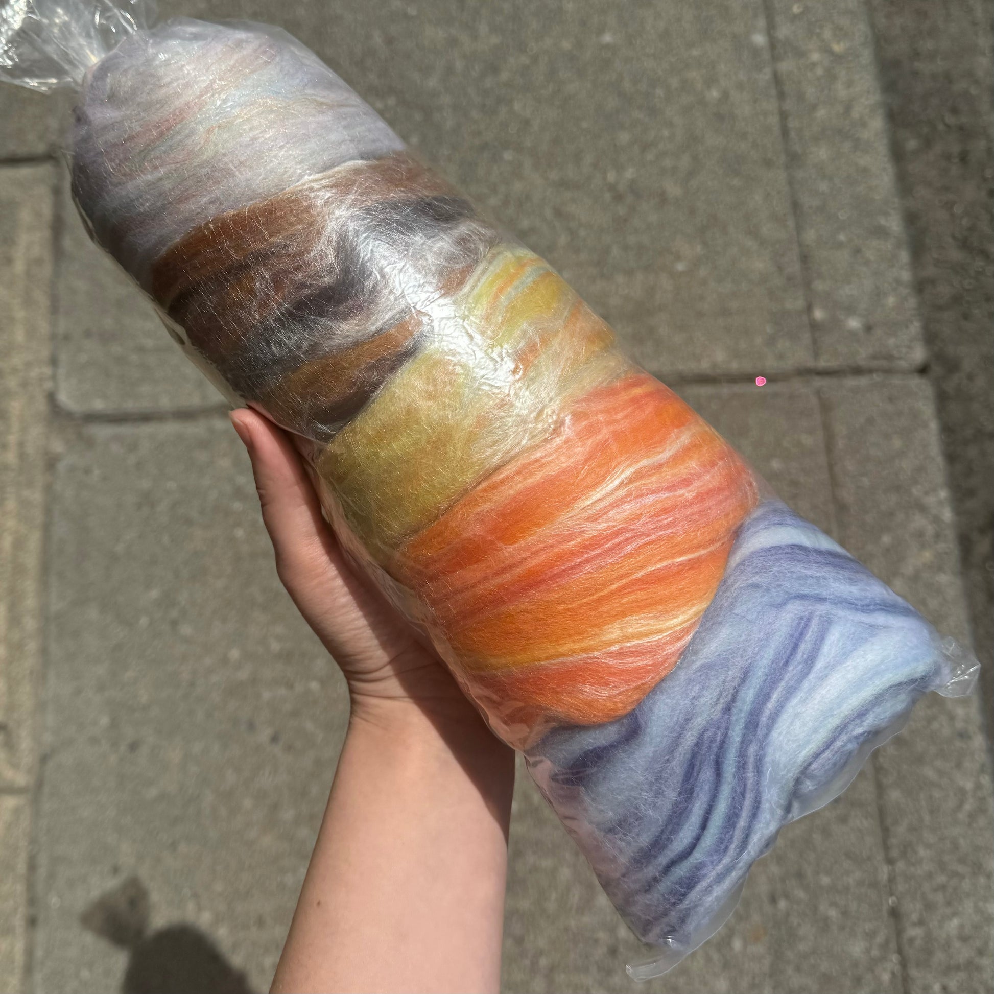 A hand is holding a large roll of colorful wool roving wrapped in clear plastic. The wool includes shades of white, brown, yellow, orange, and blue. The background shows a concrete sidewalk.