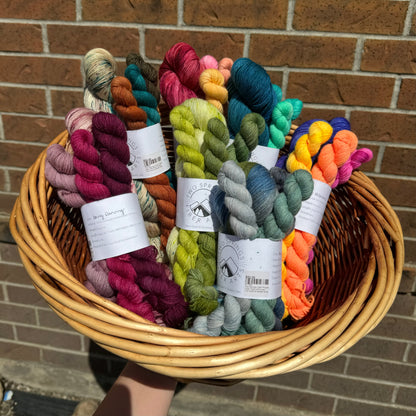 A basket filled with various colorful skeins of yarn, including shades of red, purple, green, blue, and multicolored. Each skein has a label with brand information. The basket is held outside against a brick wall background, partially in sunlight.