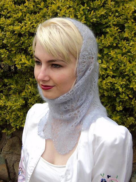 A person with short, blonde hair is wearing a light blue lace headscarf and a white garment. They are standing in front of a leafy green bush. The person is looking slightly to the left with a neutral expression.