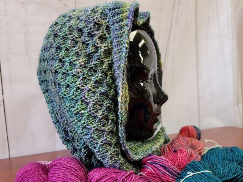 A black mannequin head is displayed wearing a multi-colored, knitted hood. The hood has a textured pattern. In the foreground, skeins of variously colored yarn are arranged next to the mannequin. The background is made of white wooden panels.