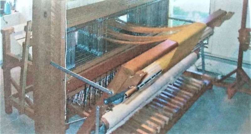 A wooden handloom with multiple threads and weaving materials is set up in a bright room. The loom is equipped with various levers and bars, with a partially woven fabric visible. The floor beneath shows supporting slats and parts of the loom structure.