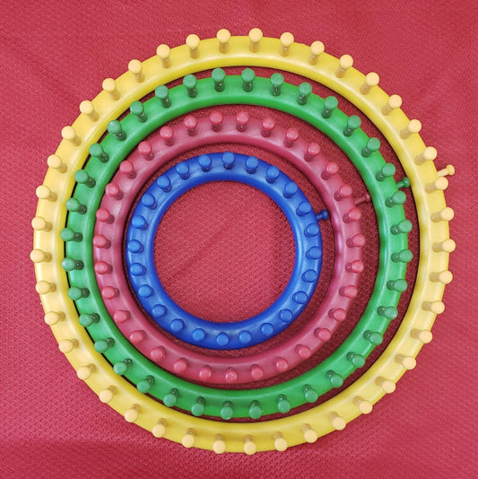 Four circular knitting looms of different sizes and colors (yellow, pink, green, blue) with evenly spaced pegs are arranged concentrically on a red, textured surface.