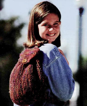 A girl is smiling at the camera while wearing a denim jacket. She is carrying a knitted backpack with a buttoned flap over her shoulder. The background is blurred, showing some trees and a pole.
