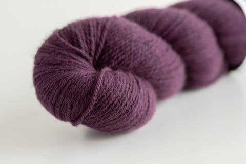 A close-up image shows a skein of deep purple yarn. The yarn is coiled neatly, highlighting its soft and slightly fuzzy texture. The photo captures the details and richness of the yarn's color and fibers.