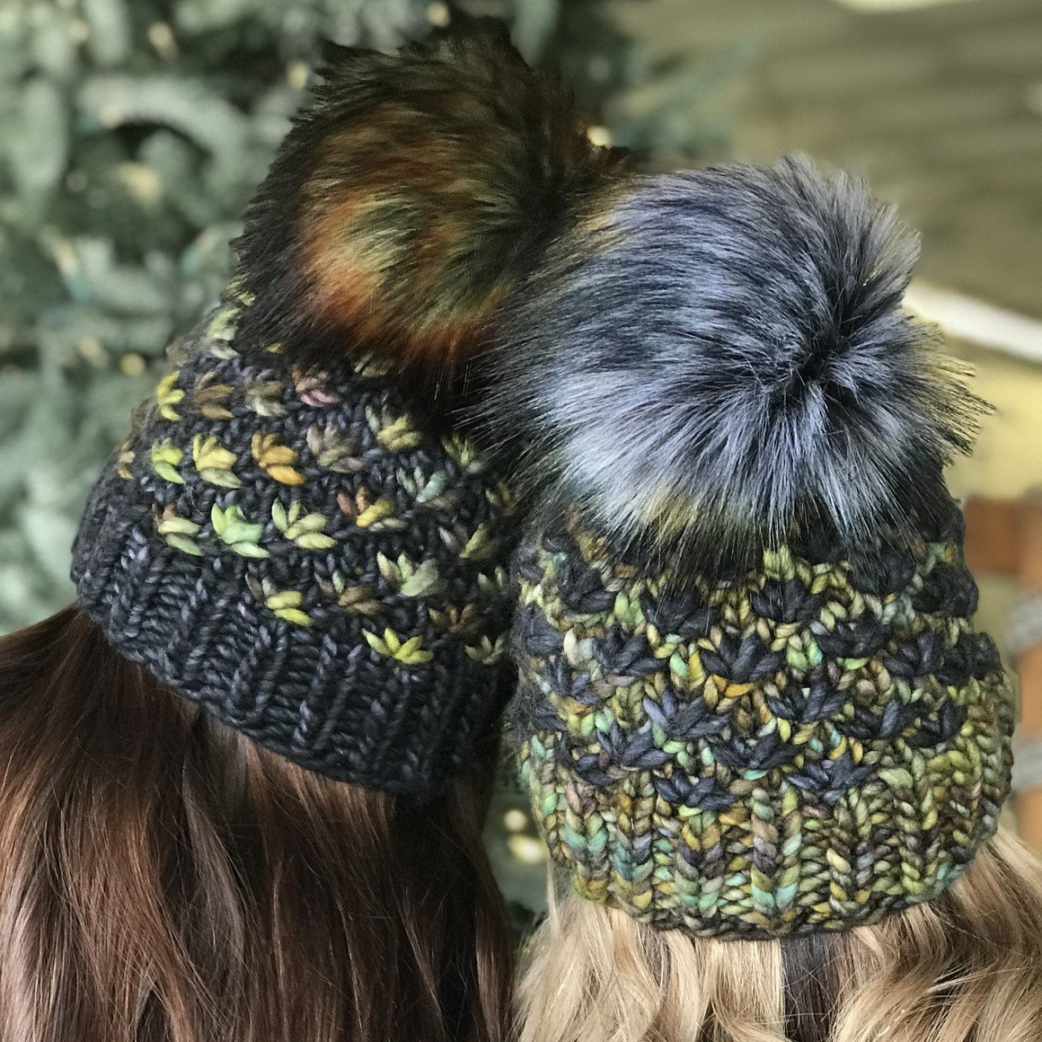 Two people, seen from the back, wearing knitted hats with large, fluffy pom-poms. The hats are made from multicolored yarn with shades of green, blue, and yellow, and feature a textured pattern. A blurred background with greenery is visible.