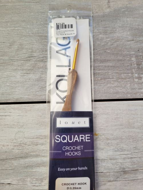 A packaged square crochet hook with a wooden handle and a gold-colored hook is displayed on a light wooden surface. The packaging includes text stating "Louet Square Crochet Hooks", "D: 3.25 mm", and "Easy on your hands". A price sticker of $13.25 is on top.