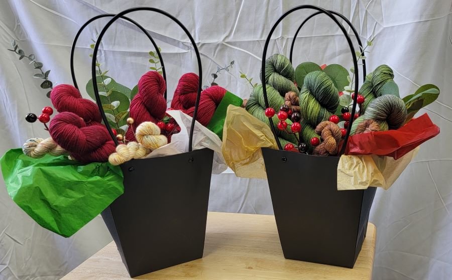 Two black gift bags containing yarn skeins and decorative elements stand on a wooden surface. Each bag has a mix of red, green, and beige yarn, accentuated with faux berries and greenery, and is lined with colorful tissue paper. A white draped background is visible.