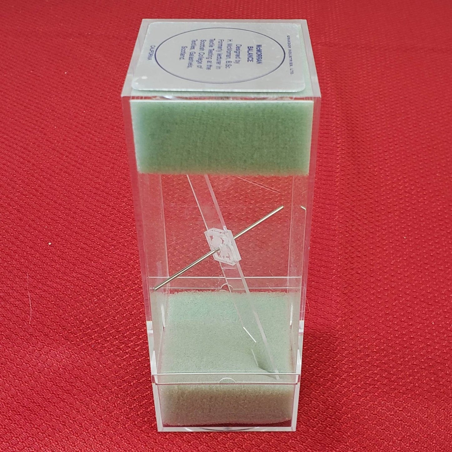 A transparent rectangular box is shown against a red textured background. The box contains green foam padding at the top and bottom, with two diagonal metal pins crisscrossing in the center, held together by a white plastic piece.