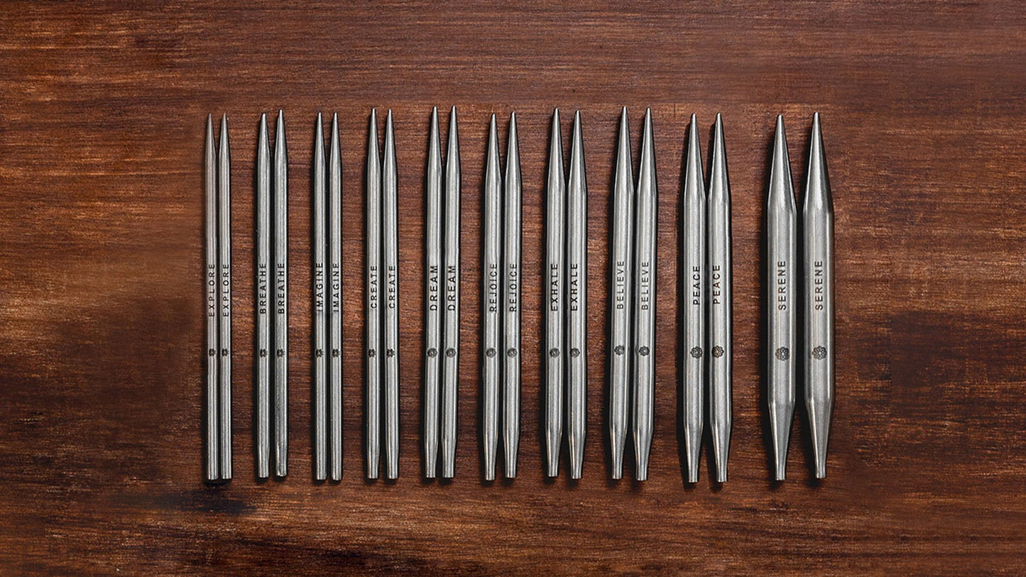 A set of metal knitting needle tips organized in a row on a wooden surface. The needles vary slightly in length and thickness, with each pair labeled with size and brand markings. The surface has a rustic brown wood finish.