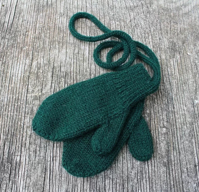 A pair of green knitted mittens connected by a string is placed on a weathered wooden surface. The mittens appear to be child-sized and have a simple, classic design.