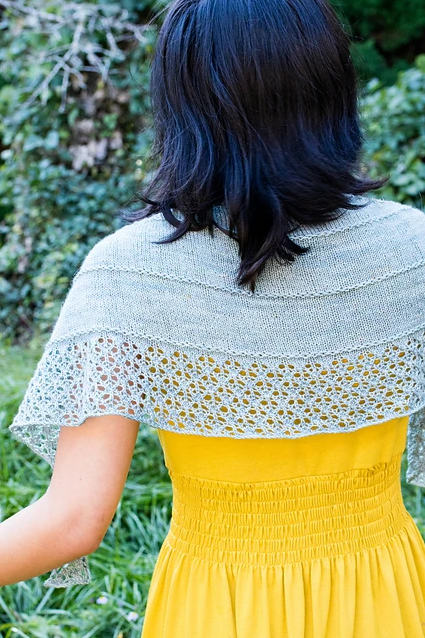 A person with shoulder-length dark hair is photographed from behind, standing outdoors. They are wearing a bright yellow dress and a light gray knit shawl. The background features green foliage and grasses.