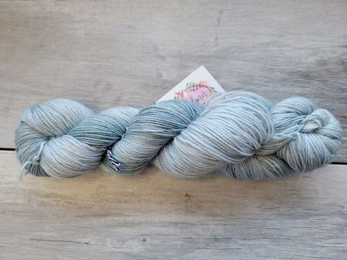 A skein of light blue and grey yarn is placed on a light wooden surface. A small tag with a floral design is attached to the yarn. The yarn appears soft and evenly spun.