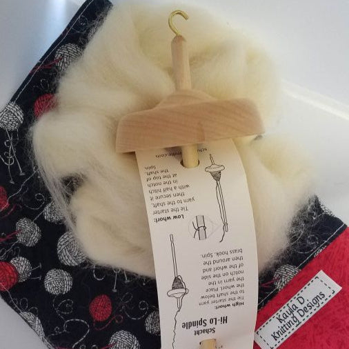 A wooden spindle with a bundle of fiber attached to it, displayed on top of a red and black patterned fabric project bag.
