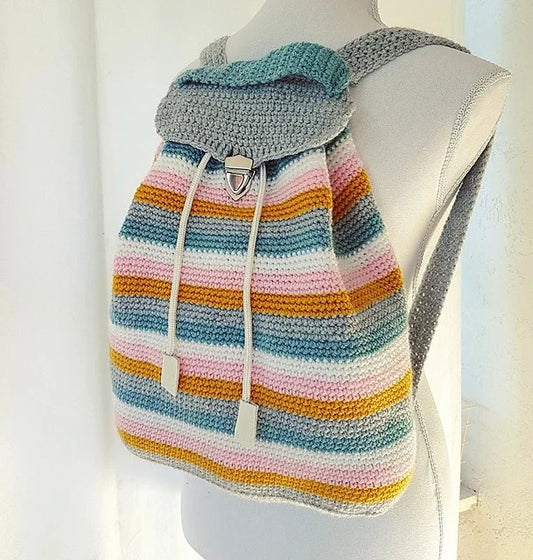 A crocheted backpack displayed on a mannequin. The backpack features horizontal stripes in various colors including teal, pink, yellow, and grey. It has grey straps, a flap closure, and two drawstrings with white tips.