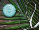 A teal retractable measuring tape with intricate white patterns and the text "SHAPES" sits coiled against a backdrop of green leaves on a dark wooden surface.