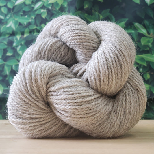 A wooden table displays multiple skeins of yarn in various shades of brown, grey, and cream. The background consists of a green, leaf-patterned fabric. The yarn is neatly arranged in rows on the tabletop.