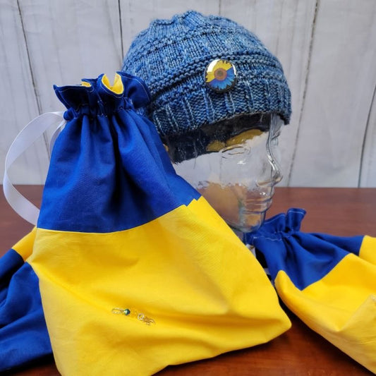 A glass mannequin head is wearing a blue knitted hat with a sunflower pin. Attached to the glass head's base are blue and yellow drawstring bags, one of which is pinned with another sunflower pin and some small metal ornaments.