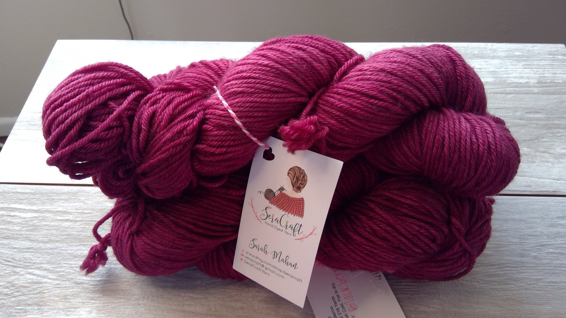 Three skeins of deep magenta yarn are displayed on a light wooden surface. Each skein has a white label attached, which features a logo with a woman knitting and reads "Sheep Crafters" and "Worsted Wool.