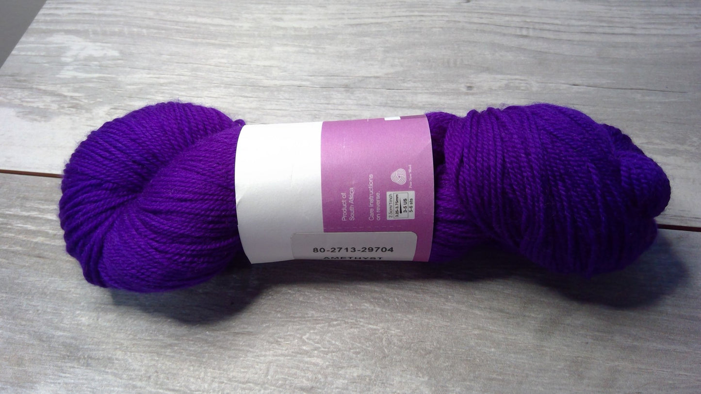 A skein of purple yarn with a white and purple label on a gray wooden surface. The yarn is coiled in a tight bundle. The label displays several barcodes and text, including the product number "80-2713-29704.