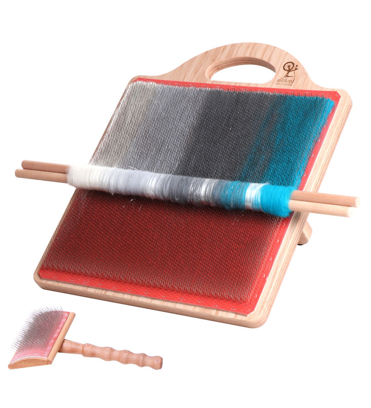 A wooden blending board with multicolored fibers arranged on its surface, including shades of white, gray, black, and blue. Two wooden dowels rest horizontally on top. A wooden-handled carding brush is positioned in front of the blending board.