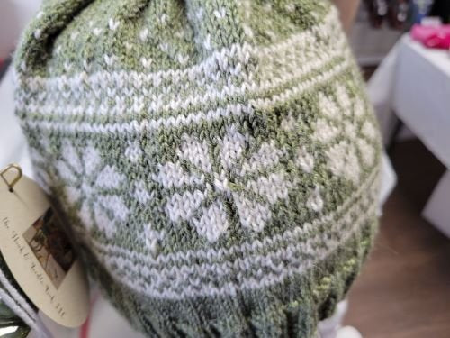 A close-up of a knitted hat featuring green and white patterns. The design includes white flower-like shapes and geometric motifs on a green background. A tag is partially visible, hanging from the side of the hat.