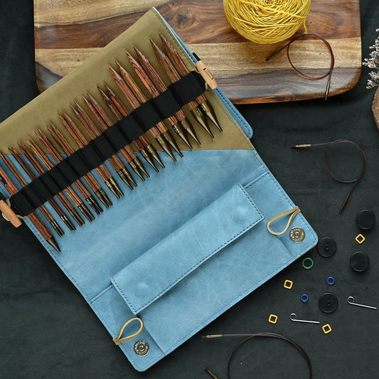 A knitting needle case is open, revealing an organized set of wooden knitting needles in slots. Scattered around it on a table are various knitting accessories, such as stitch markers, buttons, and a ball of yellow yarn. The case is blue and beige.