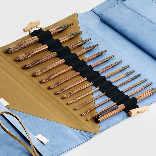 A rolled-out fabric case displays an assortment of wooden crochet hooks of various sizes. The case is blue with a brown interior and secured with elastic bands holding the hooks in place. Two ribbon ties are visible for closing the case.