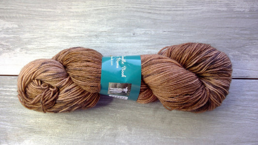 A skein of brown and beige variegated yarn lies on a textured wooden surface. The yarn has a label showing the brand name and a small picture, indicating it is a specific type or brand of yarn. The colors blend smoothly from light to dark shades.