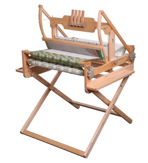 A wooden weaving loom stands on a folding wooden frame. The loom has a partially woven green and white fabric visible, with various wooden parts and levers. The overall structure appears sturdy and functional for textile creation.