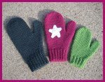 Three knitted mittens are displayed on a textured surface. One mitten is dark gray, another is pink with a white star, and the third is green. The pink and green mittens appear to be smaller in size compared to the dark gray mitten.