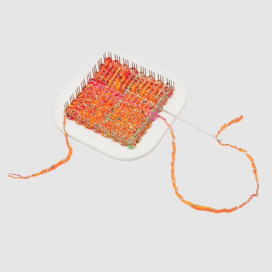 A square loom with orange, pink, and green threads is partially woven on a white plastic frame. A needle with orange thread is attached to the fabric. The loose ends of the thread extend from the weaving project.