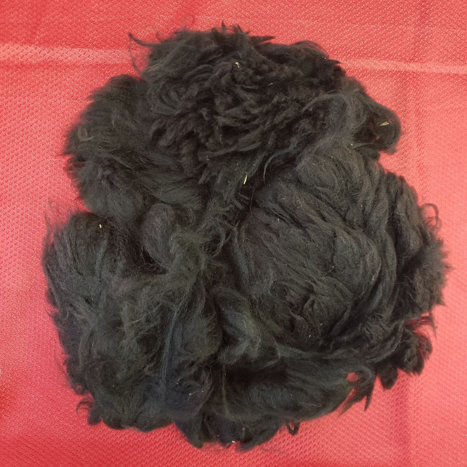 A pile of black wool is on a red fabric background. The wool appears unprocessed and fluffy, with a mix of thick and thin strands. Some small bits of debris are visible in the wool.