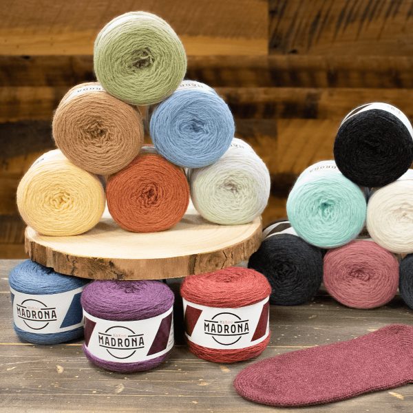 A display of assorted yarn balls in various colors. Two small pyramids of yarn are on wooden surfaces; nearby, several more yarn balls are arranged alongside a knitted swatch. Each yarn ball is labeled "Madrona." The background is rustic wood.